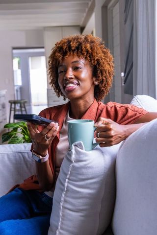 African american woman relaxing on sofa holding teal mug and remote, cozy living room
