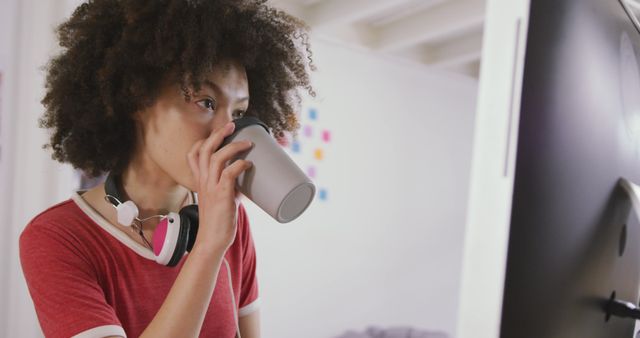 Young woman focused on her work, drinking coffee at her desk with headphones around her neck. Perfect for illustrating remote work, home office setup, and modern work-life balance. Suitable for articles on productivity, work from home tips, or tech lifestyle.