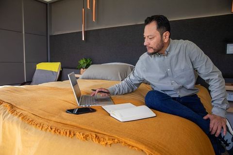 Mature asian man working remotely on laptop from bedroom with notebook and phone