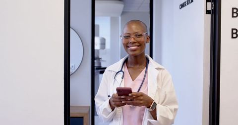 Smiling clinician holding smartphone checking messages wearing lab coat over pink scrubs