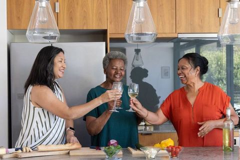 Multicultural mature women toasting wine at kitchen island, cozy home entertaining