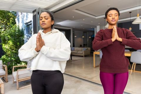 Indian and african american women meditating in prayer pose by open sliding glass door