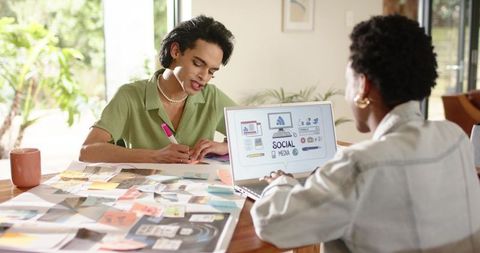 Diverse duo brainstorming social media campaign at home desk with moodboard and laptop