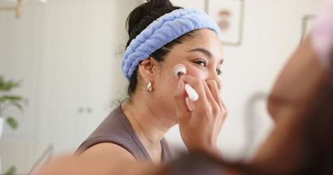 Woman applying makeup at home vanity wearing blue headband and gold hoop earrings