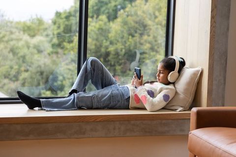 Asian teen girl lounging on window seat listening with headphones and using phone