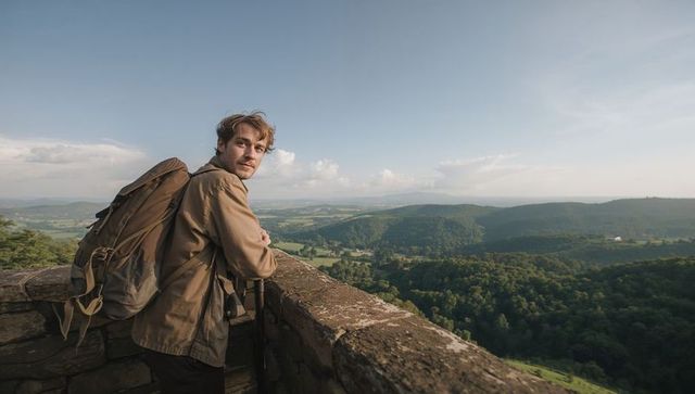 Hiker leaning on stone wall overlooking rolling green valley and distant forested hills