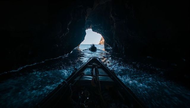 Bow of boat cutting through dark sea cave toward sunlit ocean opening, coastal voyage