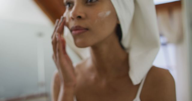 Biracial woman wearing towel on head applying cream on her face. domestic life, spending quality free time relaxing at home.