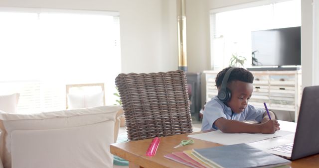 Young boy wearing headphones is focused on studies while sitting at a desk at home. Various school supplies are scattered on the desk. This image can be used for websites, articles, and advertisements related to online education, homeschooling, and virtual learning platforms.