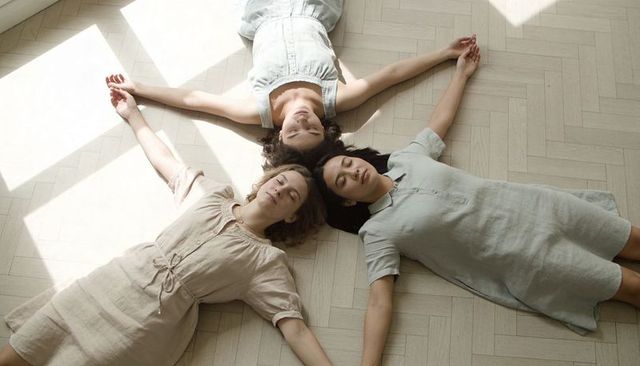 Women lying in star formation on sunlit parquet floor wearing linen dresses, minimal calm