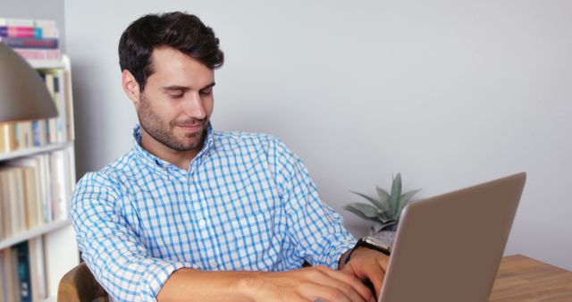 A young Caucasian man is focused on his laptop while working at a desk, with copy space. His casual attire suggests he may be working from home or in a relaxed office environment.