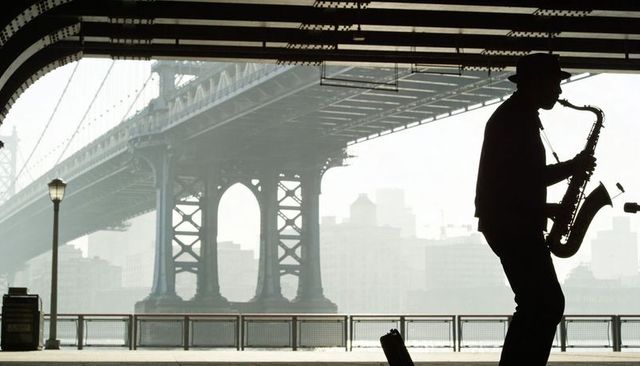Busker playing tenor sax under manhattan bridge, silhouette backlit on waterfront