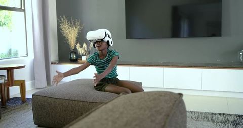 African american girl playing virtual reality with headset on ottoman in living room
