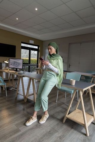 Woman wearing green hijab leaning on trestle table checking phone in modern coworking