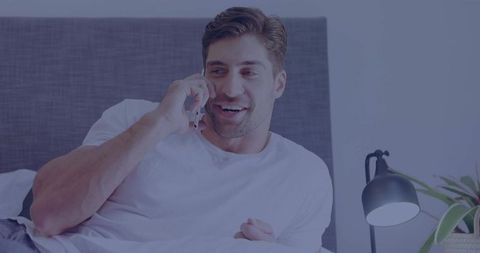 Smiling man relaxing on bed while talking on smartphone in cozy modern bedroom