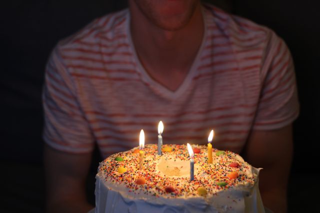 A person wearing a red and white striped shirt sitting in front of a cake with lit candles. The cake is decorated with colorful sprinkles, making the moment festive. Perfect for use in birthday-themed content, celebrating milestones, or advertising party supplies and decorations.