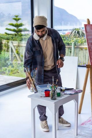 Indian artist arranging brushes in bright seaside studio wearing beanie and headphones