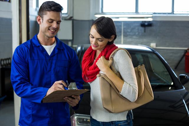 Mechanic in blue uniform showing checklist to female customer in auto repair garage. Customer holding handbag and wearing red scarf, looking at checklist. Ideal for illustrating car maintenance services, customer service in auto repair shops, and professional consultations in automotive industry.