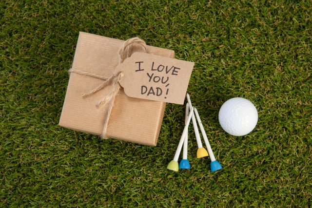 Brown gift box with twine and handwritten 'I Love You, Dad!' note on grass next to golf ball and colorful tees. Ideal for Father's Day promotions, gift ideas, and celebration themes. Perfect for advertisements, greeting cards, and social media posts.