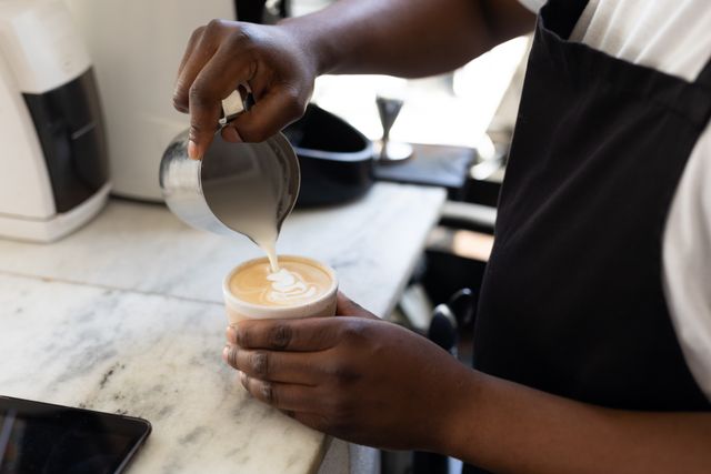 This image shows a young African American barista skillfully pouring milk into a coffee cup, creating latte art. Ideal for use in articles or advertisements related to coffee culture, barista training, cafes, and the service industry. It can also be used in social media posts promoting coffee shops or barista skills.