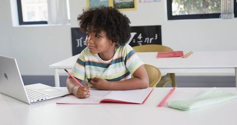 African American School-age Boy Studying with Laptop Writing Notes in Classroom Math