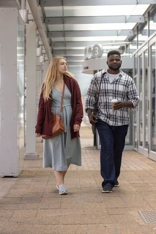 Diverse couple walking through airport concourse under glass canopy carrying coffee and shoulder bag