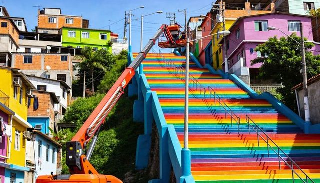 Workers repairing handrail on rainbow-painted hillside staircase using orange boom lift