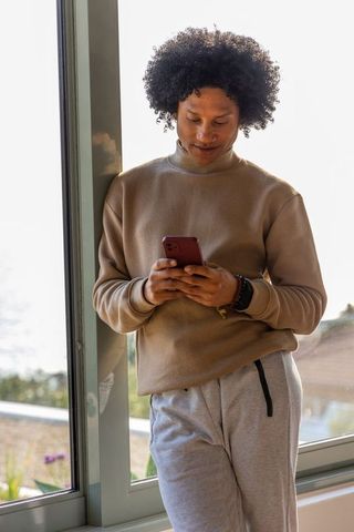 African american man leaning by glass door texting on red smartphone in soft light