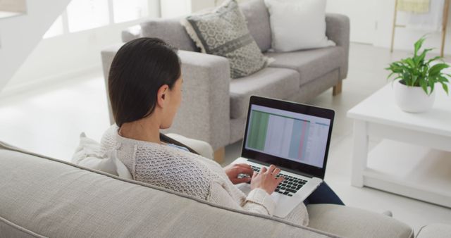 A woman is working from home, sitting on a sofa in her living room, analyzing data on her laptop screen. The modern living room setting, natural light, and casual attire suggest a comfortable and efficient work-from-home environment. This image could be used for articles or posts about remote work, home office setups, work-life balance, modern living rooms, home decor, and technology in daily life.