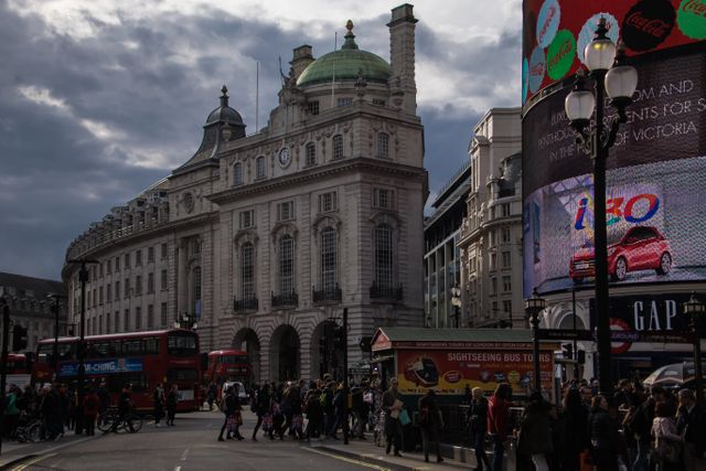 People walking and sightseeing in Piccadilly Circus, London. Iconic double-decker buses, historic buildings, and bright advertisements in the plaza. Useful for travel guides, websites, and articles about London, tourism, city life, and historic landmarks.