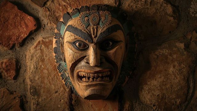 Carved wooden ceremonial mask with braided cords, faded pigments hanging on rustic stone wall