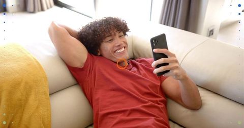 Young man relaxing on couch smiling while using smartphone in bright living room