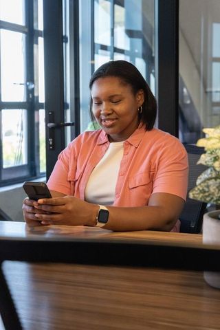 African american woman using phone at office desk wearing smartwatch, smiling and focused
