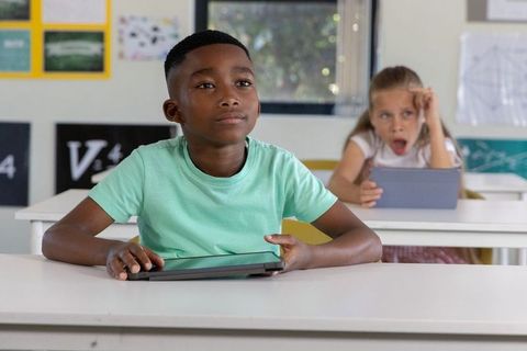 Young student concentrating with tablet in classroom while classmate yawning