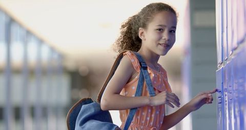 Smiling schoolgirl opening locker with blue backpack in hallway