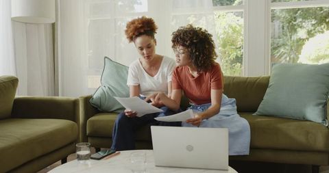 Two women reviewing documents on sofa with laptop for home office collaboration