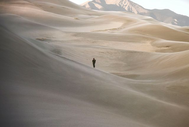 Solitary figure walking across expansive desert dunes with mountains in the background. This scene conveys themes of isolation, adventure, and the beauty of untouched nature. Perfect for travel blogs, adventure stories, and promotional materials emphasizing tranquility, vast landscapes or the spirit of exploration.