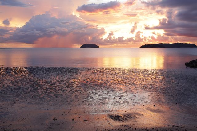 Dramatic sunset over serene beach, calm waters reflecting the vibrant sky colors. Islands silhouetted against the horizon, clouds adding depth to the picturesque scene. Perfect for travel websites, nature magazines, meditation apps, or interior décor emphasizing tranquility and natural beauty.