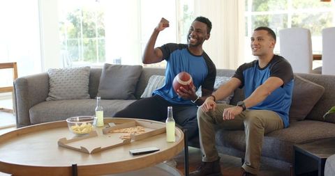 Two friends cheering while watching football at home holding football pizza and drinks