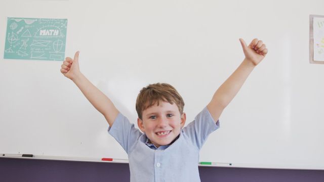 Boy standing in front of a whiteboard, raising thumbs excitedly in a classroom. Useful for illustrating positive school environment, promoting educational content, or advertising academic programs.