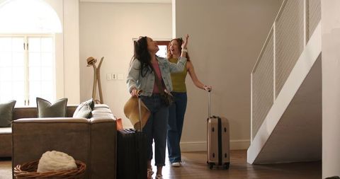 Female friends arriving at cozy home pulling wheeled suitcases while holding straw hat