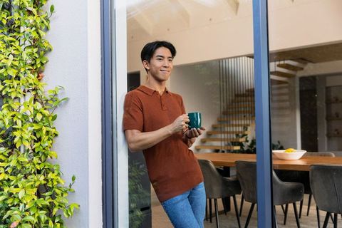 Adult asian man standing in glass doorway holding green mug, modern dining and staircase