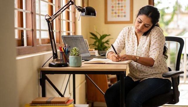 Woman working from home writing notes while talking on phone at sunlit minimalist desk