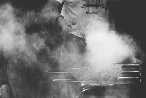Person grilling with tongs over smoky charcoal grill in black and white lifestyle scene