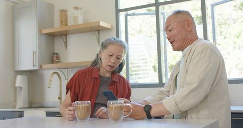 Asian senior couple sharing smartphone at bright modern kitchen island with coffee mugs