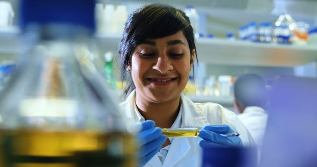 Young female scientist in lab coat and gloves smiling while examining sample in test tube in laboratory setting. Focus on scientific research and discovery. Ideal for content related to scientific research, innovation, medical advancements, chemistry studies, or educational materials.