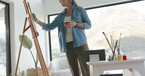 Mature woman painting at easel in sunlit lakeside studio holding cup and brush
