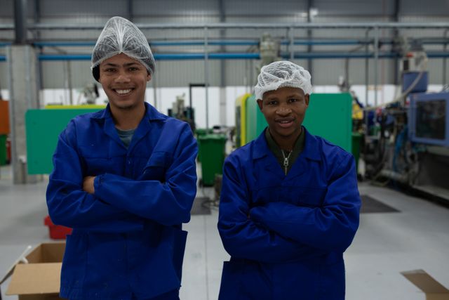 Portrait of a biracial and an African American male worker working in a busy factory warehouse wearing hair nets and blue overalls standing with arms crossed looking to camera and smiling