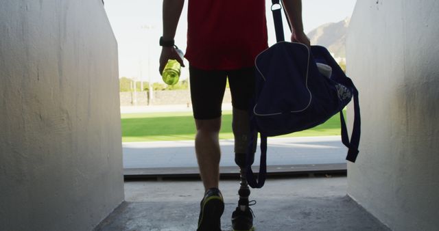 Person with prosthetic leg walking towards a sports field holding a sports bag and water bottle. Great for topics related to sports inclusivity, determination, adaptive athletes, motivation, fitness.