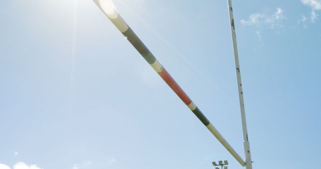 A view from below captures a high jump bar against a clear blue sky, with copy space. Sunlight flares from the side, adding a dynamic element to the potential of athletic achievement in track and field events.